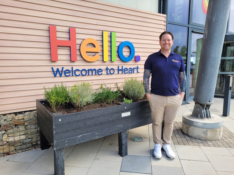 Chris May standing beside raised herb planter in front of colourful “Hello Welcome to Heart” sign at Tesco HQ