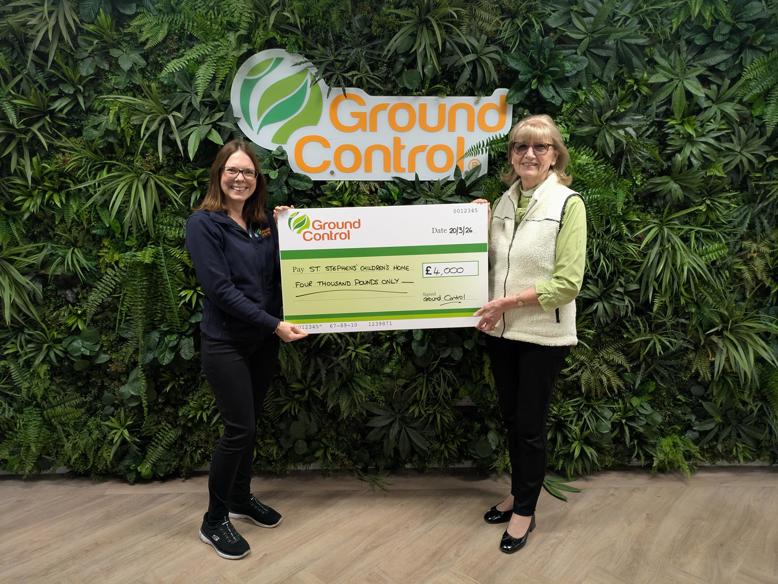 Two people hold a large charity cheque in front of a green wall with the Ground Control logo.