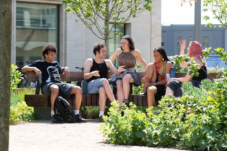 People seated on a wooden bench in a landscaped courtyard, surrounded by planting beds, trees and pathways outside a modern building.