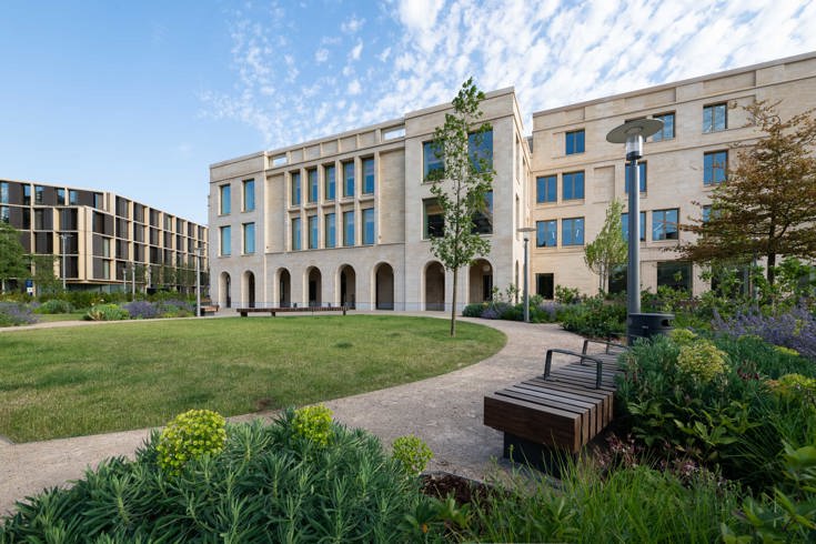Modern stone-clad office building surrounding a landscaped courtyard with curved pathways, lawn, benches, young trees and flowering plants under a blue sky.
