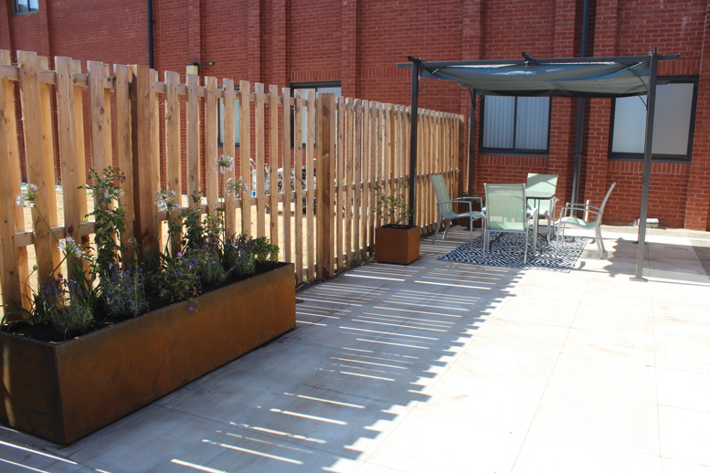 Outdoor courtyard with raised planters, timber fencing, and a shaded seating area with table and chairs beside a red brick building.