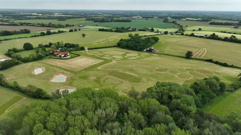 Aerial view of a rural landscape showing grassland fields, woodland, ponds and farm buildings, with large mown patterns visible across the countryside.
