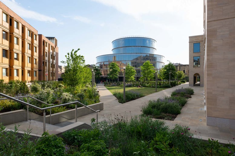 Landscaped university courtyard with planted borders, pathways and steps leading towards a modern circular glass building, surrounded by contemporary stone and brick buildings.