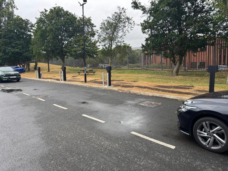 Electric vehicle charging bays along a roadside with charging bollards, parked cars and landscaped surroundings