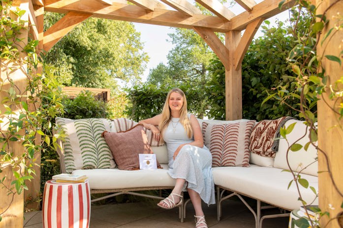 Sophie sitting on cushioned outdoor seating beneath a wooden garden pergola surrounded by greenery.